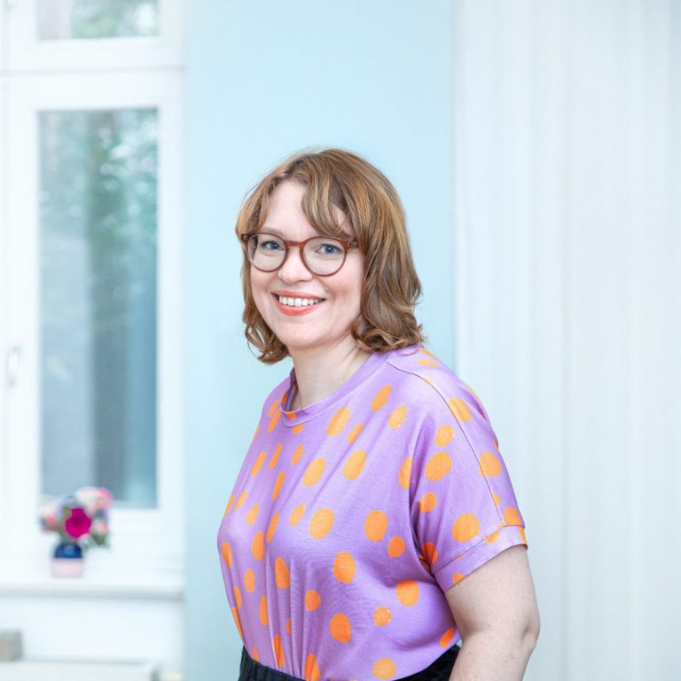 Lächelnde Frau in blauer Jacke sitzt auf einem Stuhl mit Buch in der Hand.