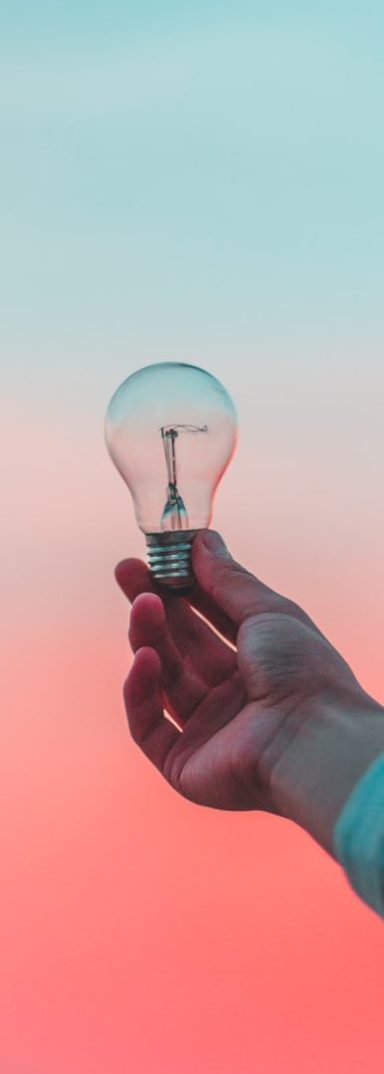 Eine Hand hält eine Glühbirne vor einem pastellfarbenen Hintergrund.