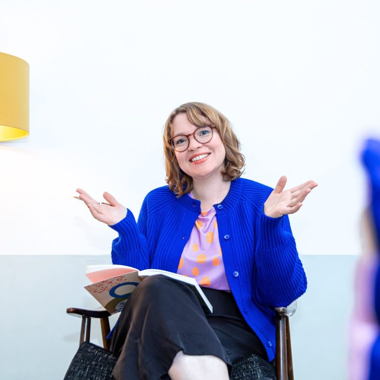 Junge Frau mit Brille, blauer Bluse und rosa Cardigan vor hellem Hintergrund.