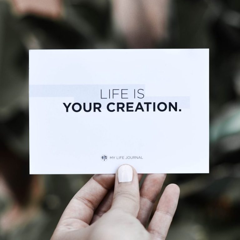 Hand hält eine Karte mit dem Text: „LIFE IS YOUR CREATION.“ in klarer Schrift.