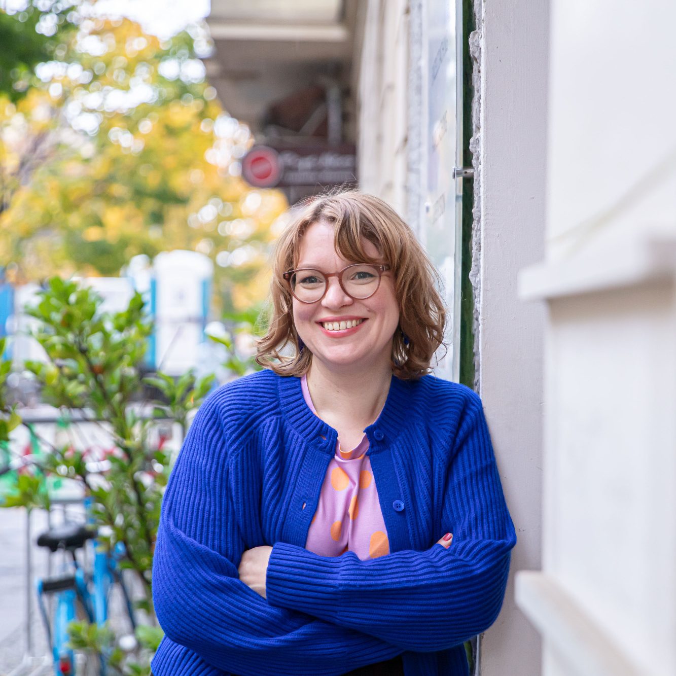 Lächelnde Frau in blauer Jacke steht vor einer Wand, umgeben von Pflanzen.